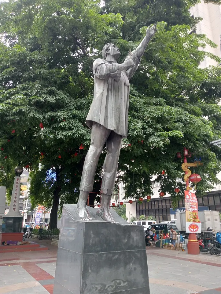 Gray metal statue of Lorenzo Ruiz on a tall stone pedestal in a city plaza, one arm raised toward the sky, with trees and shops behind