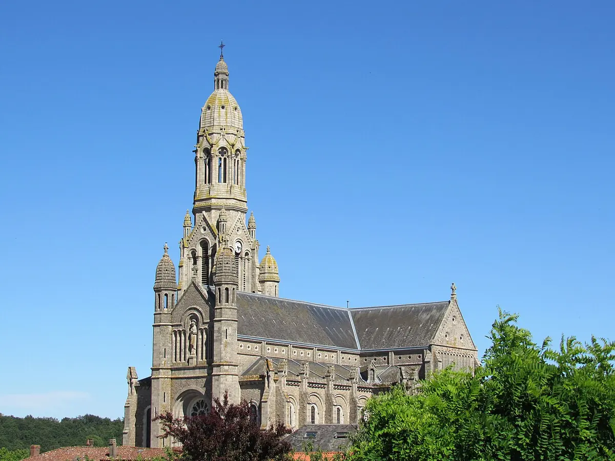 A large gray stone church with a tall domed bell tower rising above trees under a clear blue sky