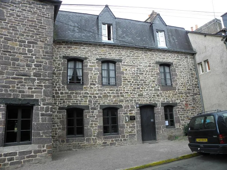 A two-story gray stone town house with slate roof and dormer windows on a narrow street in Montfort-sur-Meu