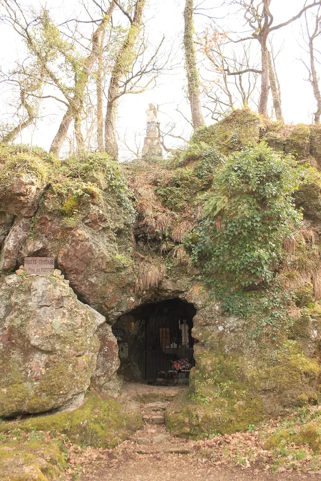 A small cave chapel with a wooden grille set into a mossy rock outcrop among bare trees, with a statue on top of the rock