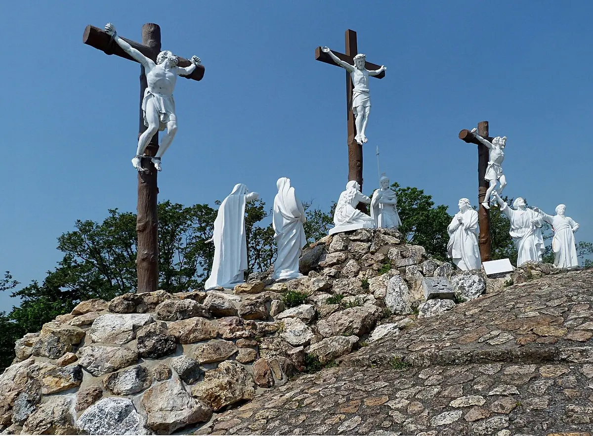 Three tall crosses with white crucified figures on a rocky mound against a blue sky, with white statues of Mary, John and the holy women below
