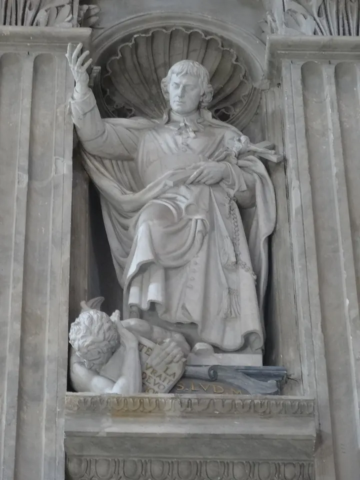 A white marble statue in a shell-topped niche showing Montfort raising one hand and holding a crucifix and rosary in the other, above a crouching winged demon that clutches a book inscribed Traité de la vraie dévotion