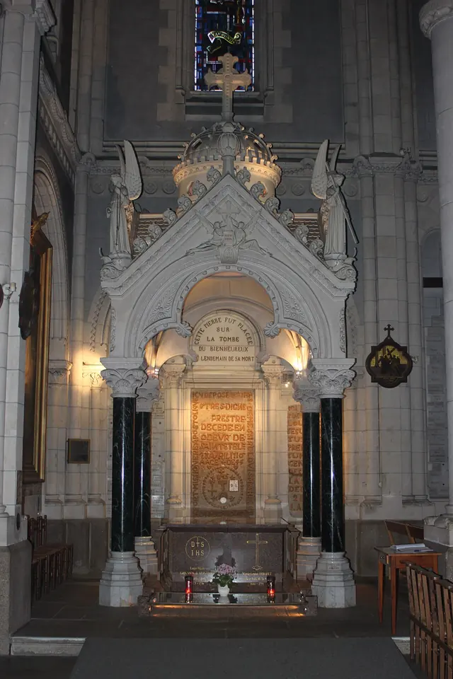 A white marble shrine with dark columns and a carved Gothic canopy built against a church wall, framing an inscribed tomb slab, with candles in front