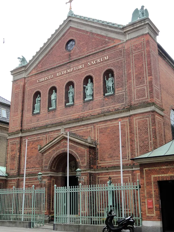 Red-brick church facade with a triangular gable, the inscription Christo Redemptori Sacrum and a row of pale statues of saints in niches over the door