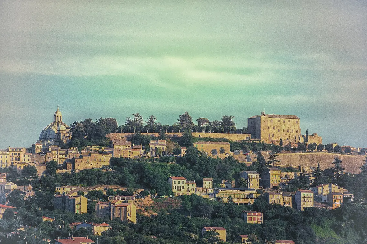 Hill town of ochre houses rising to a fortress and a domed cathedral on the summit, under a pale green-blue sky