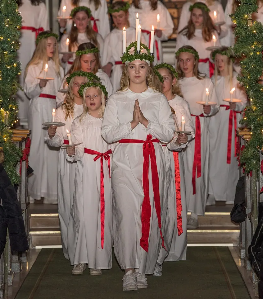 Una niña con corona de velas encendidas y túnica blanca con faja roja encabeza una procesión de niñas vestidas de blanco con velas por una iglesia de Suecia