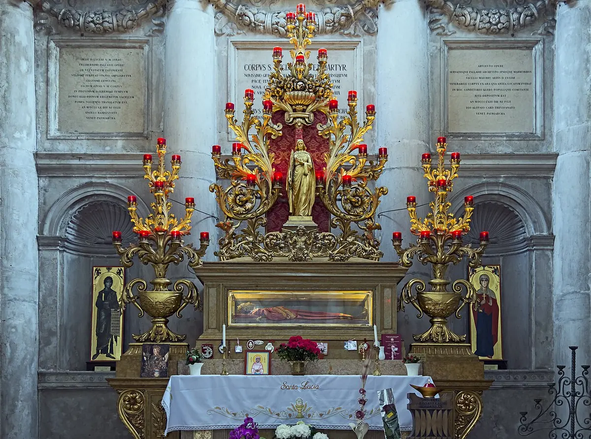Capilla de mármol con altar dorado y altos candelabros; bajo el altar, una urna de cristal guarda el cuerpo de Santa Lucía con vestido rojo
