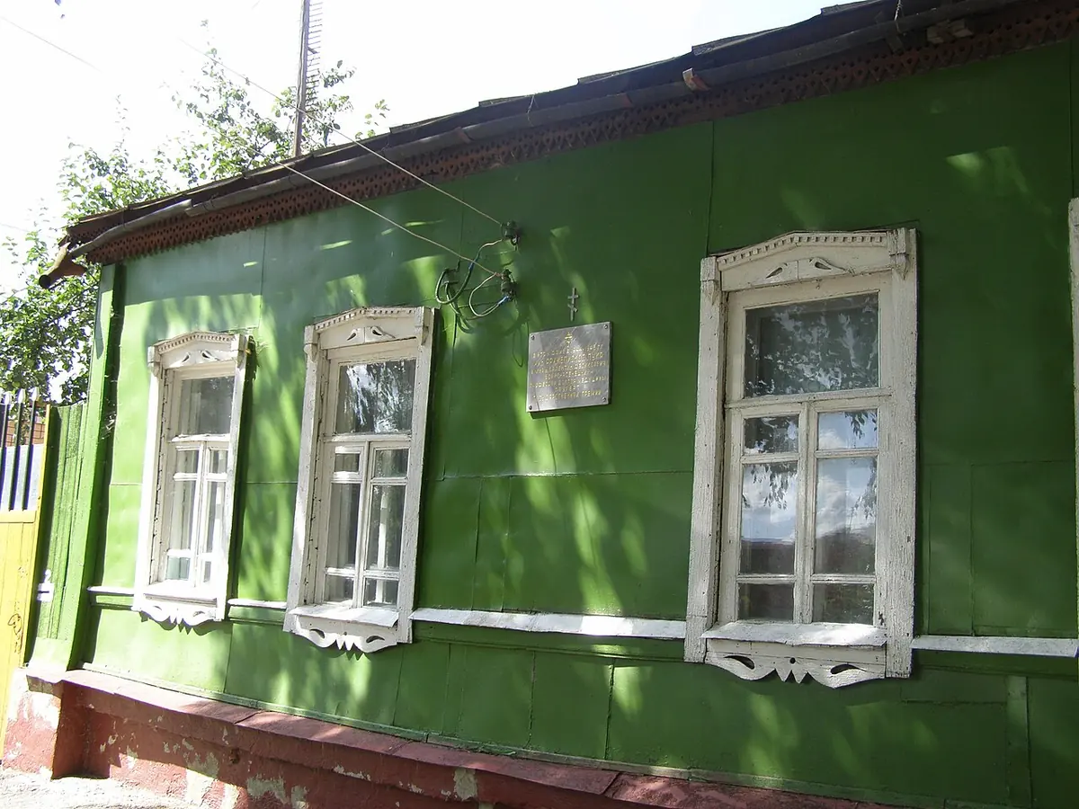 A green wooden one-story house with white carved window frames and a memorial plaque on the wall