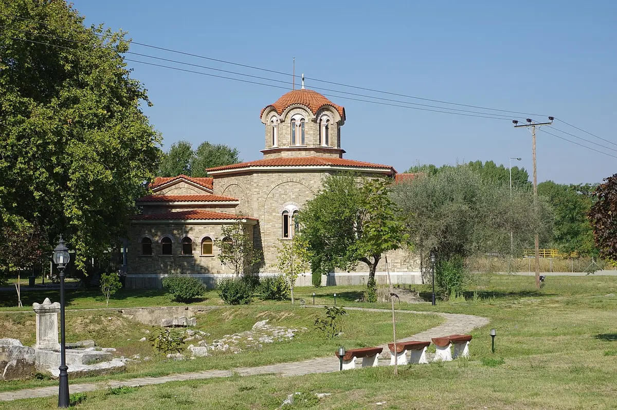Small stone church with a red-tiled dome and a lawn with ancient stone fragments in front, under a blue sky