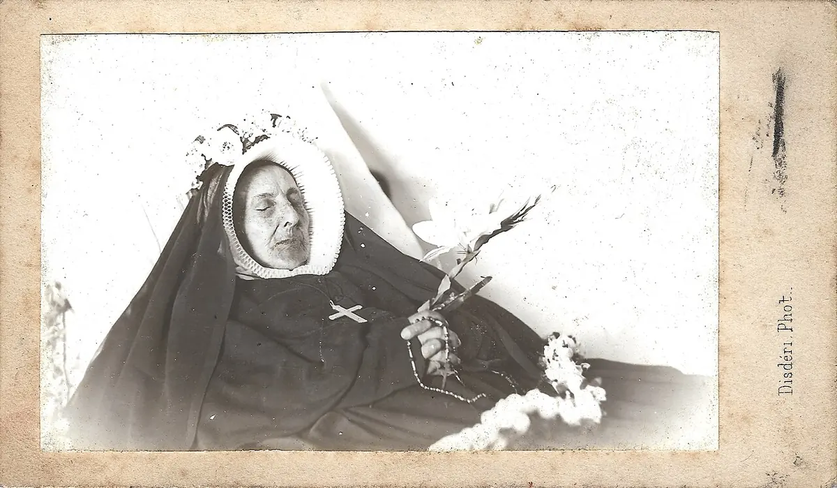 Old photograph of an elderly nun lying in death in her black habit and white bonnet, with a crucifix on her breast and lilies in her hands