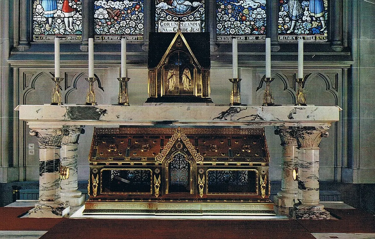 A marble altar with gilded Gothic shrine beneath it and a row of tall candles, before stained-glass windows in a chapel in Jette