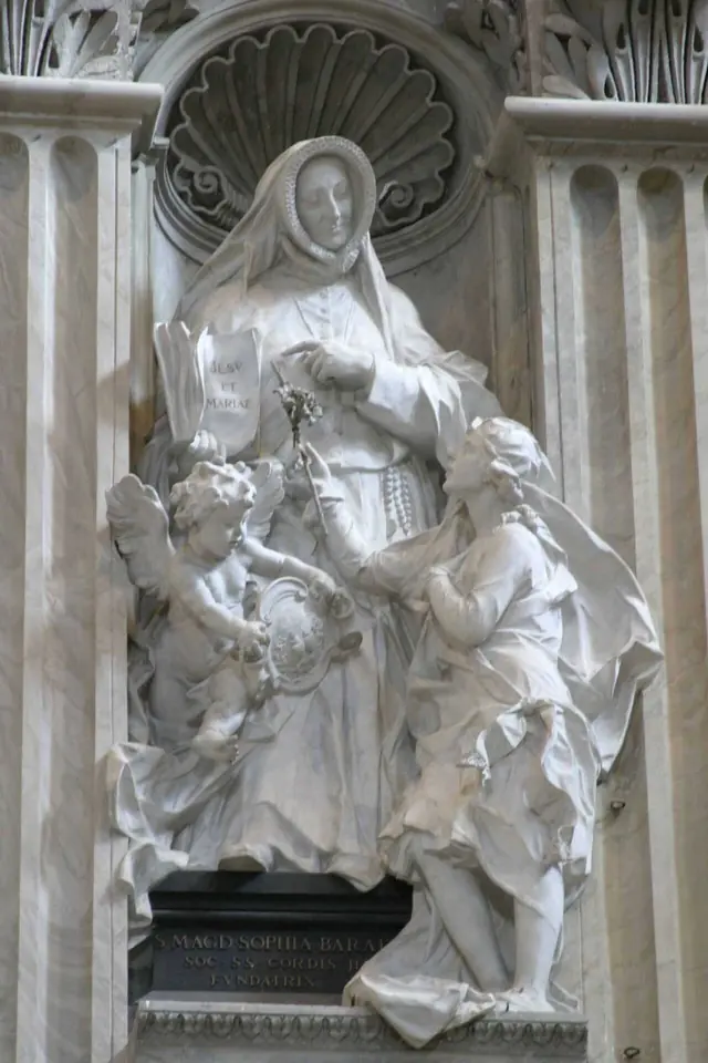 White marble statue in a niche of a nun holding a crucifix, with two children at her side, one looking up at her