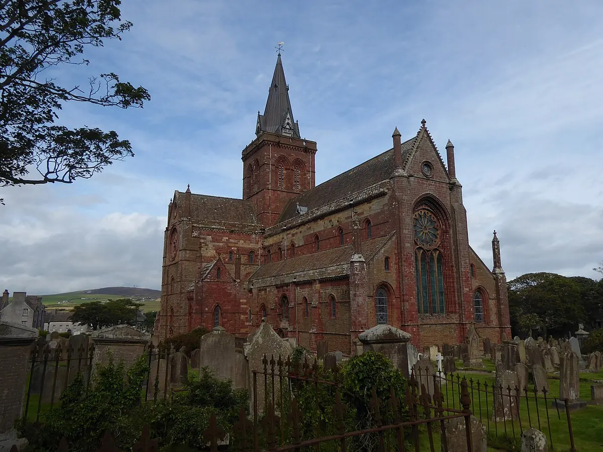 A red sandstone Romanesque cathedral with a tall spire over the crossing and a round rose window, behind a churchyard of old gravestones