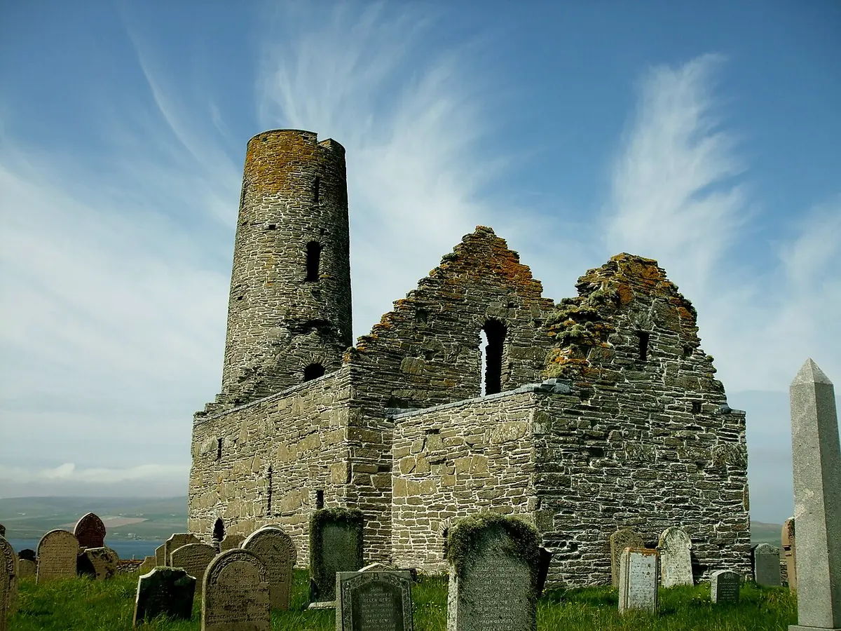 A roofless medieval stone church with a tall round tower at its west end, standing among gravestones under a blue sky with streaks of cloud