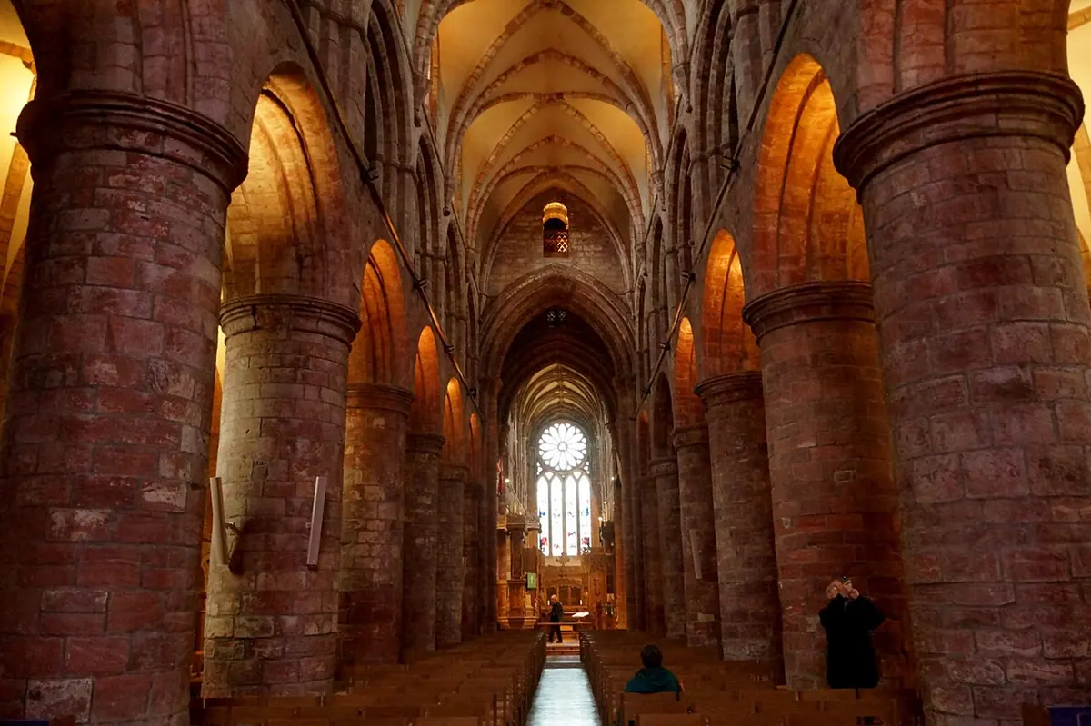 A long nave of massive round red and yellow sandstone pillars and round arches, lit warmly, leading to a rose window at the far end