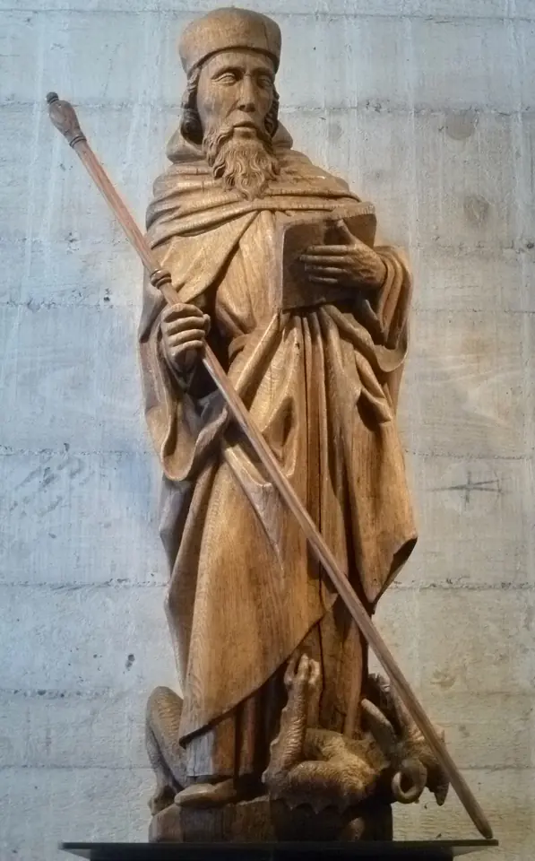 An oak statue of a bearded monk in a cap and long habit, holding a book and a long staff that pierces a dragon at his feet