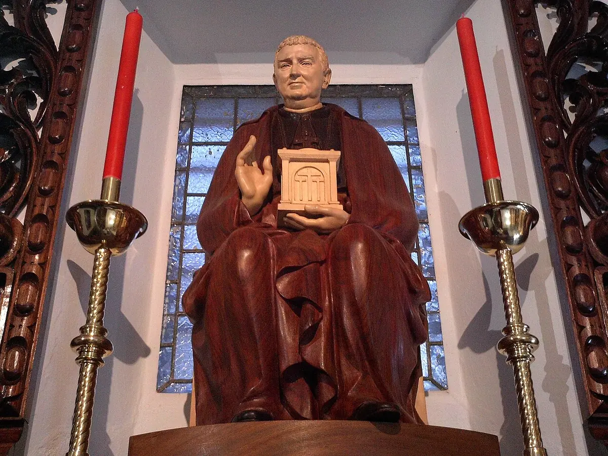 Painted wooden statue of a seated bishop in a red cape holding a small cream-colored tabernacle, between two tall red candles in a niche