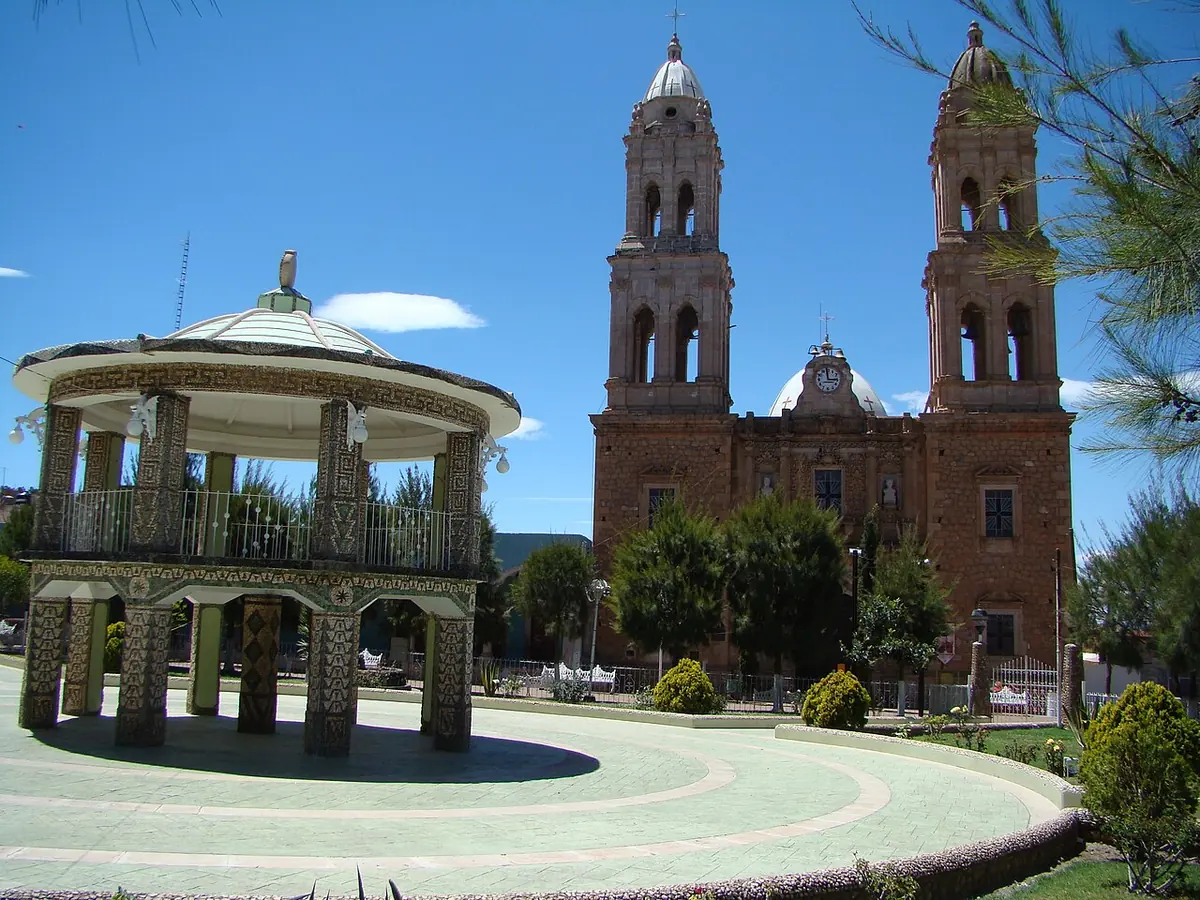 A pink stone church with two tall bell towers behind a round tiled bandstand in a sunny town square