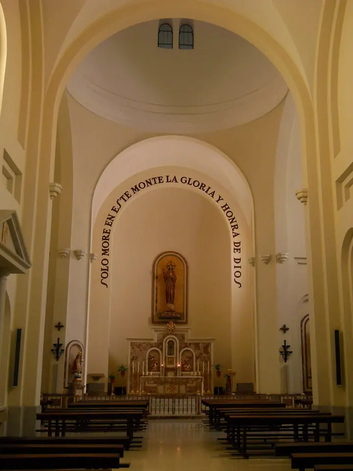 A bright, simple church nave with pale walls and a domed sanctuary, its arch painted with a Spanish verse around a small image above the altar