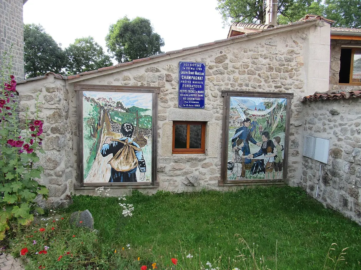 Low stone farmhouse wall with two large painted panels showing scenes of a priest and children, a blue plaque between them and flowers in front