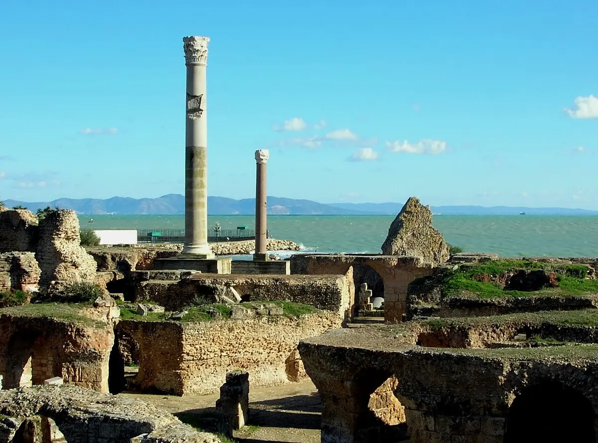 Roman ruins of brick and stone vaults beside the sea, with a single tall marble column standing against a blue sky and mountains across the bay