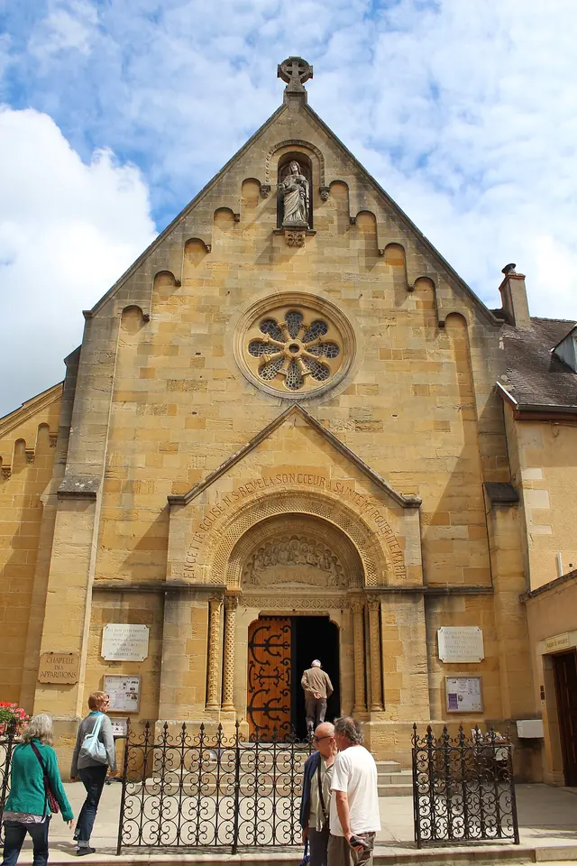 The tan stone Romanesque-style facade of the Visitation chapel in Paray-le-Monial, with a rose window, a statue in a niche and visitors at the doorway