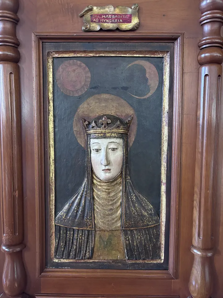 Carved and painted wooden relief of a nun's face framed by a dark veil and a small gold crown, set in a wooden frame with a Latin name plaque