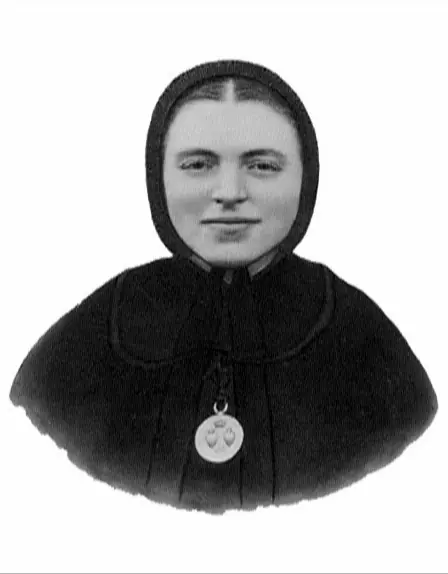 Black-and-white photograph of a young nun with a round face and a slight smile in a black bonnet and cape, wearing a round medal with two hearts