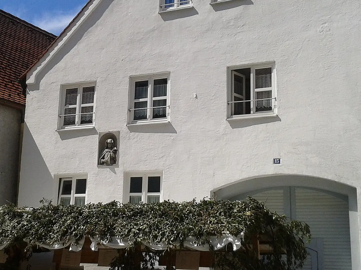 White-plastered three-story town house with small square windows, a small relief image of a saint set into the facade, and a vine-covered pergola at street level