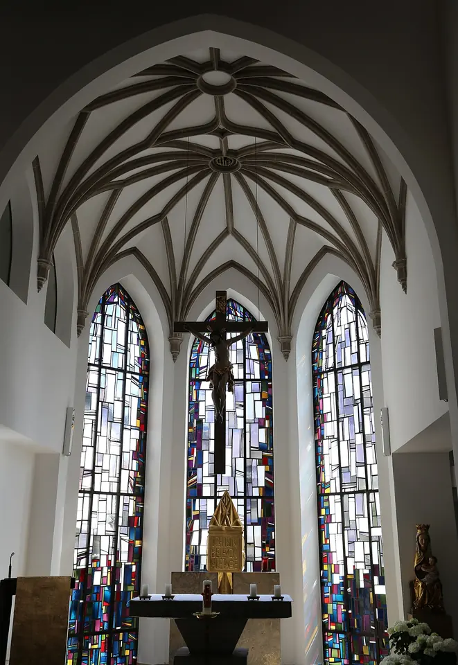 White church sanctuary with a ribbed Gothic vault, a large crucifix hanging before three tall windows of colored glass, and a simple altar