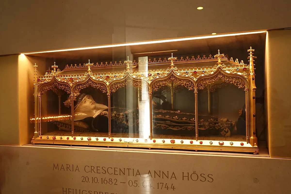 Gilded Gothic-style glass reliquary shrine lit from within, holding the clothed remains of a saint, set into a wall niche above a stone plaque with her name and dates