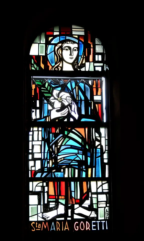 Modern stained-glass window of a girl in a blue robe holding a white lily and a green palm, with the words "St. Maria Goretti" below