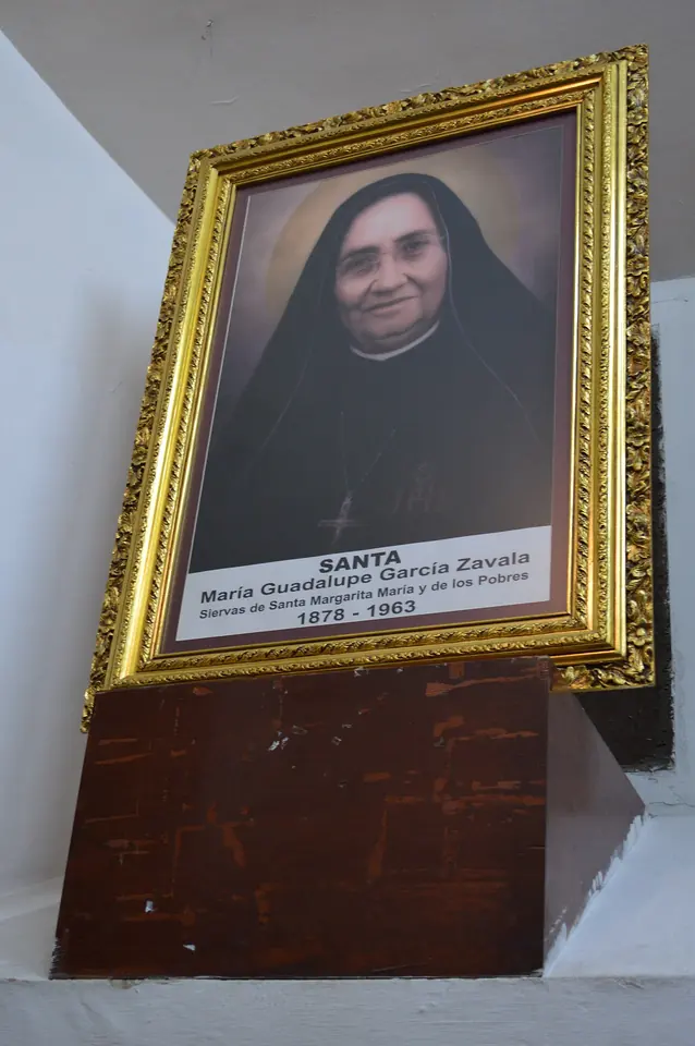 A gilt-framed portrait of a smiling nun in a black veil, labeled Santa María Guadalupe García Zavala, 1878 to 1963, on a wooden stand in a church