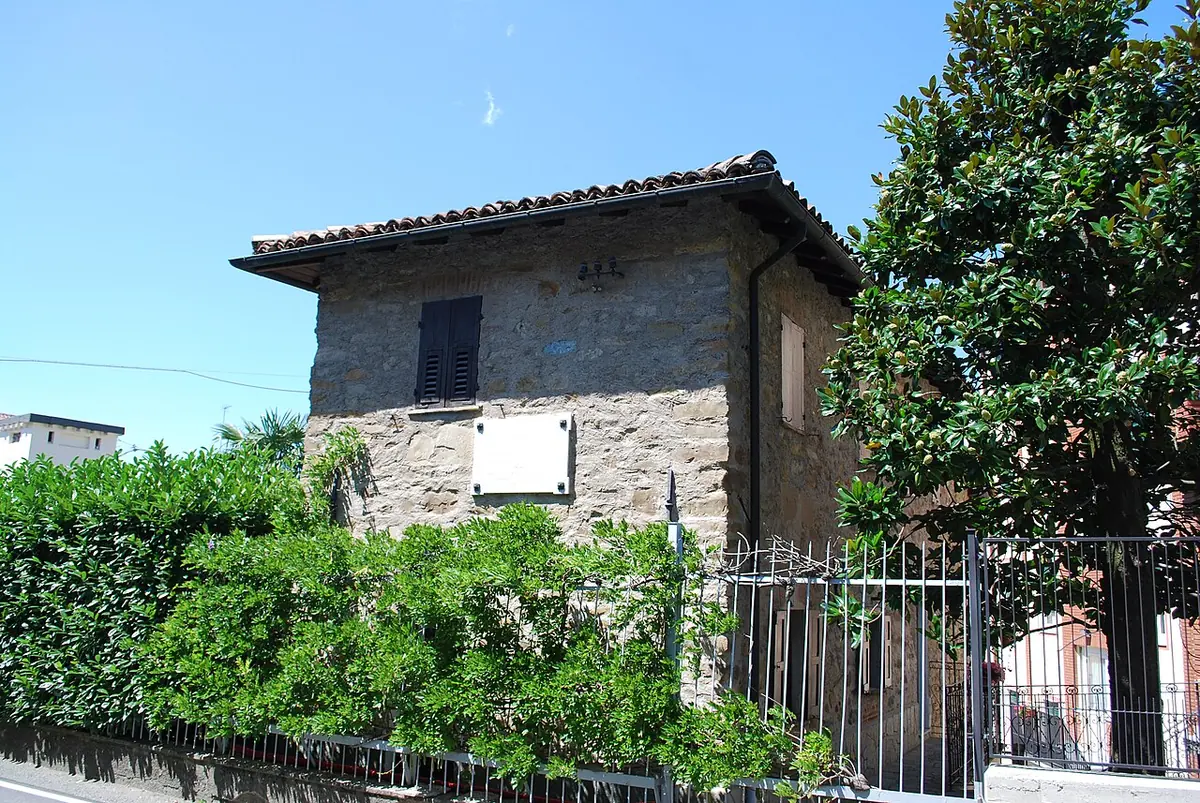 A small old stone house with a tiled roof and a white plaque on its wall, behind an iron fence and green shrubs