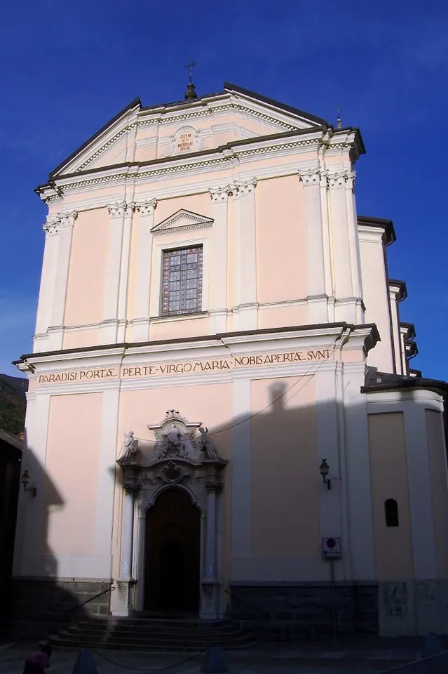 The pale pink and cream facade of a Baroque parish church with a carved stone doorway and a Latin inscription, under a deep blue sky