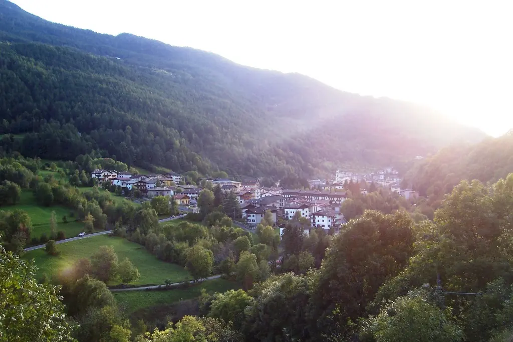 A mountain village of stone houses in a green alpine valley below steep wooded slopes, lit by the low sun