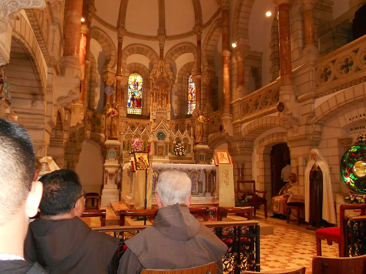 Friars and pilgrims seated in an ornate chapel with stone arches and a statue of a Carmelite nun beside the altar