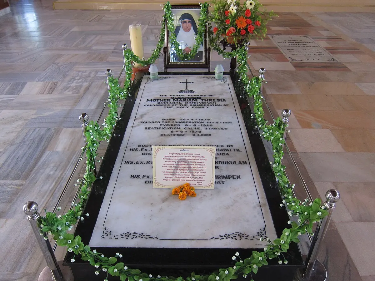 Long marble tomb slab framed by a polished wood border and a garland of green leaves, with an engraved inscription, a small framed portrait and candles at its head