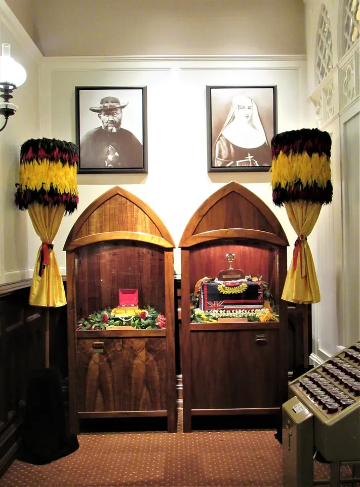 Two arched koa-wood reliquary cases side by side under framed photographs of St. Damien and St. Marianne, flanked by red and yellow feather standards