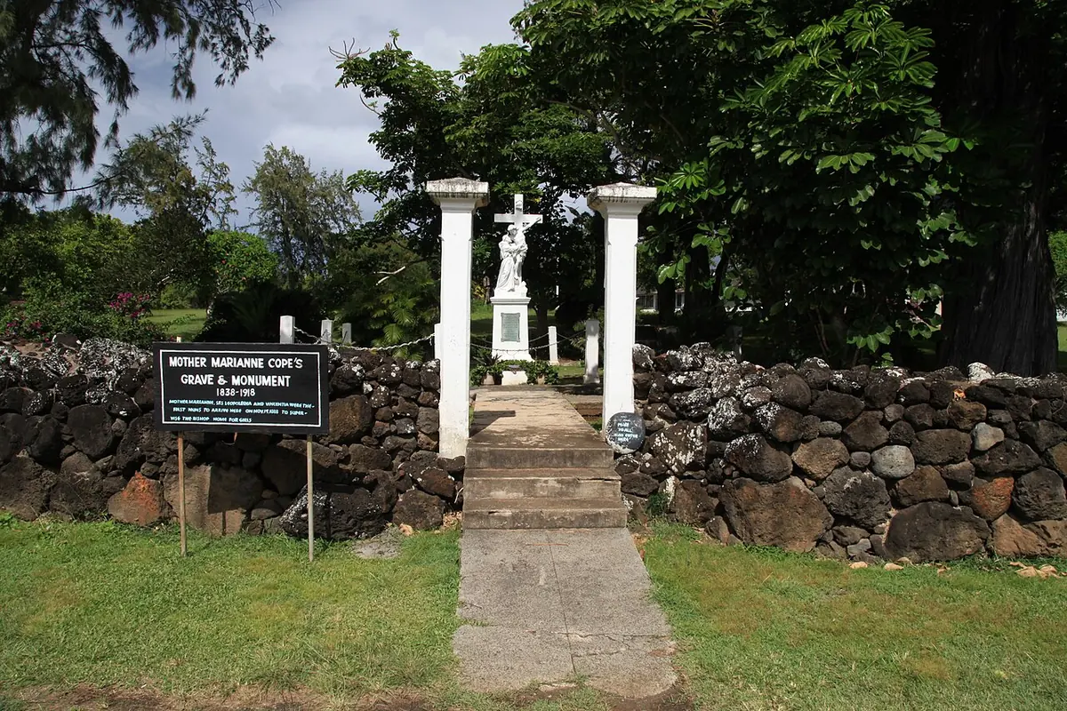 A stone path leading through a low lava-rock wall to a white statue between two white pillars, with a sign reading Mother Marianne Cope's Grave and Monument