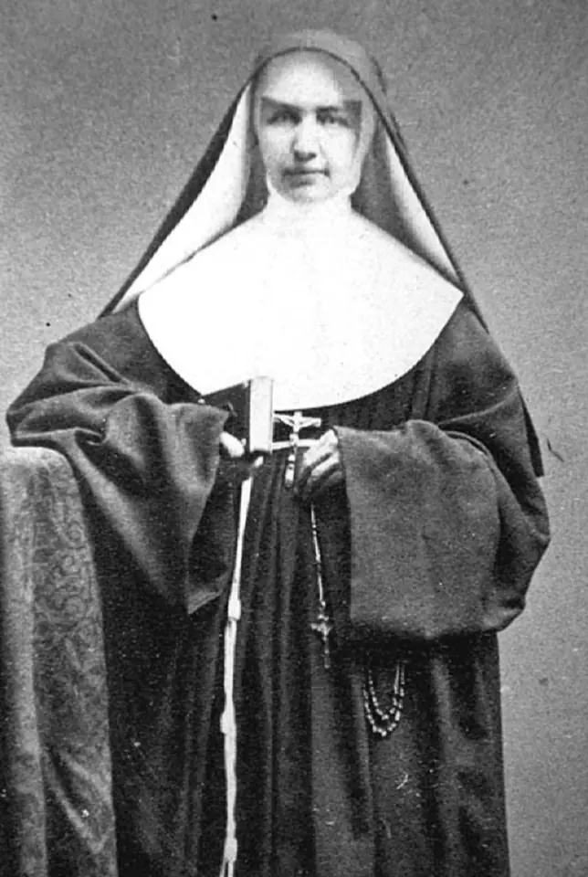 Black-and-white photograph of Mother Marianne in a black Franciscan habit and white wimple, standing beside a chair and holding a book, with a rosary and crucifix at her side