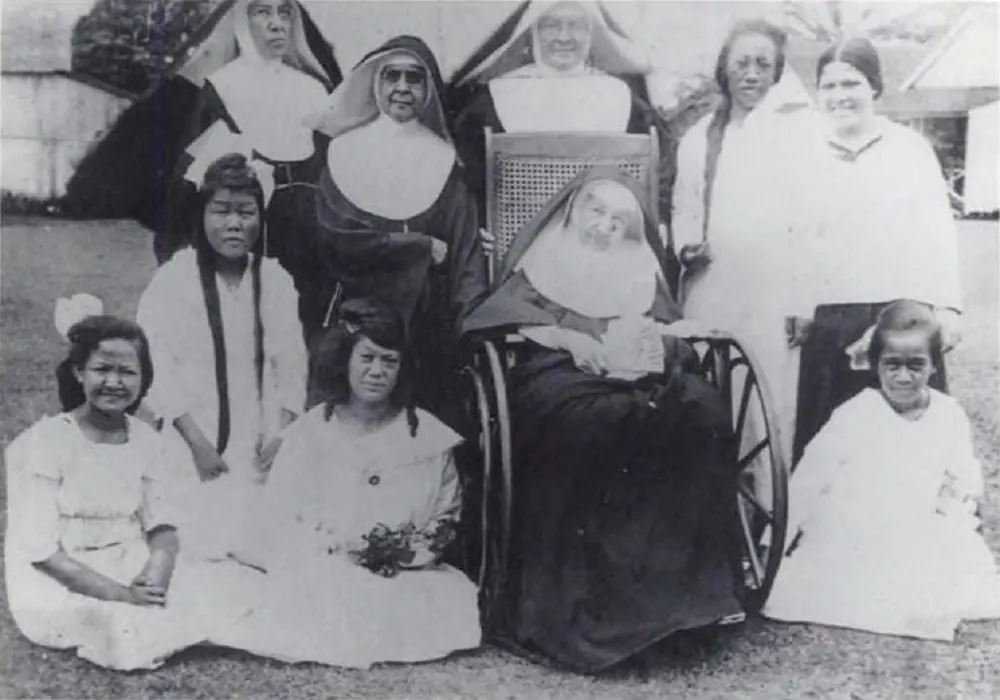 Black-and-white group photograph of an elderly Mother Marianne in a wicker wheelchair, surrounded by Franciscan sisters and Hawaiian girls and women in white dresses