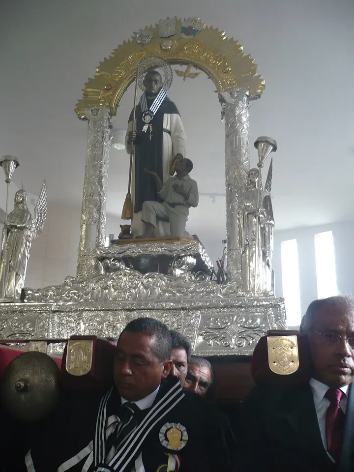 Statue of St. Martin with a broom and a kneeling figure at his side, on an ornate silver platform under a silver arch, carried by men in dark suits