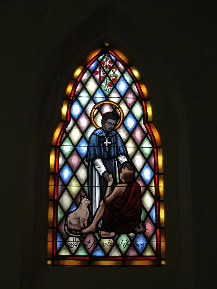 Pointed stained glass window of St. Martin in a black cape over a white habit, reaching down to a sick man in a red cloak, with a cat at their feet