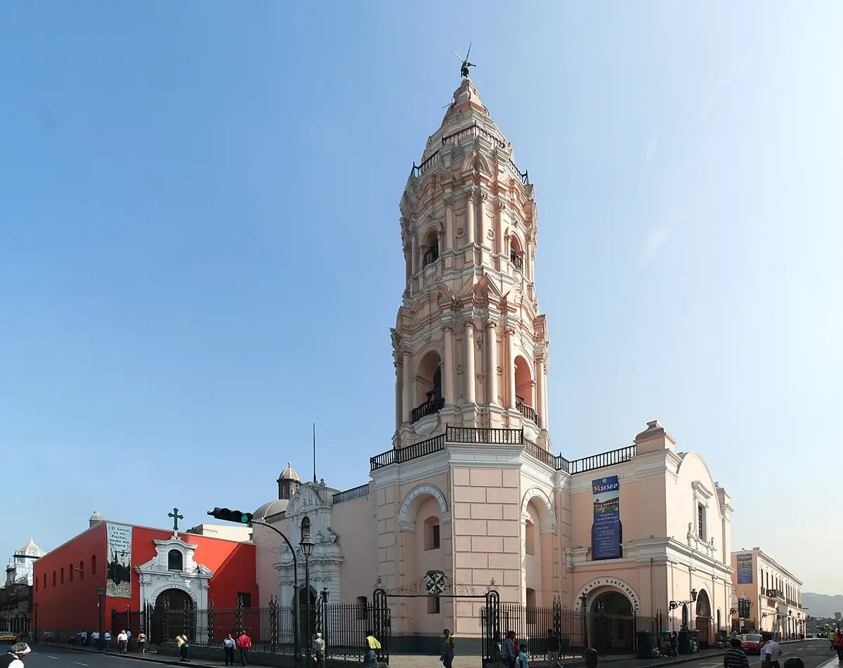Pink-and-white colonial church with a tall tiered bell tower on a sunny street corner in central Lima
