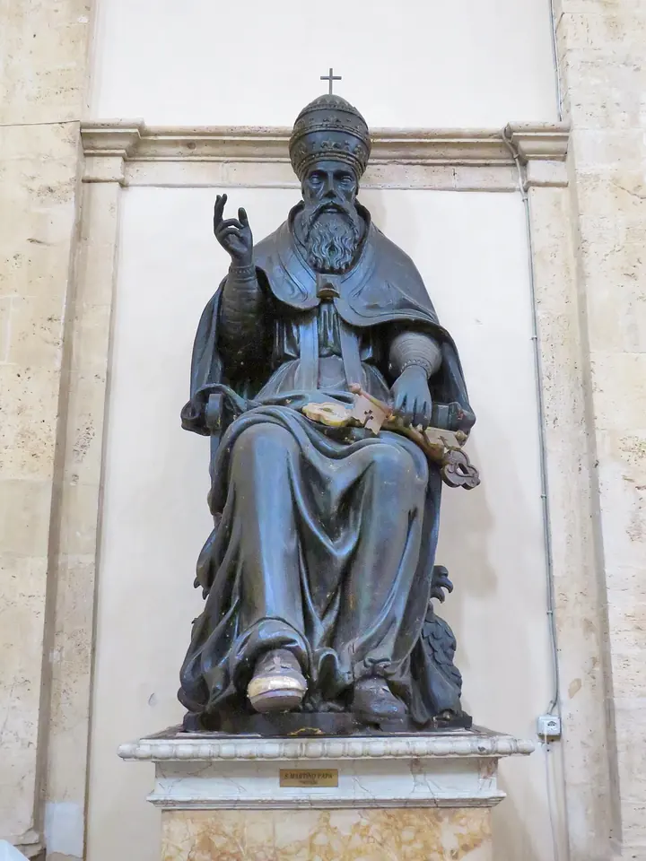 A dark bronze statue of a seated bearded pope in a tiara, raising his right hand in blessing and holding a book on his knee, in a white stone niche