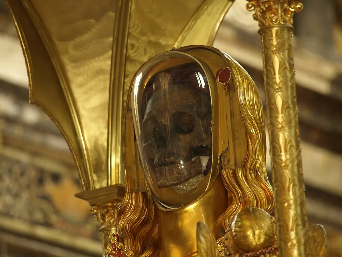 A gilded reliquary bust with long golden hair and a glass face through which a dark skull can be seen