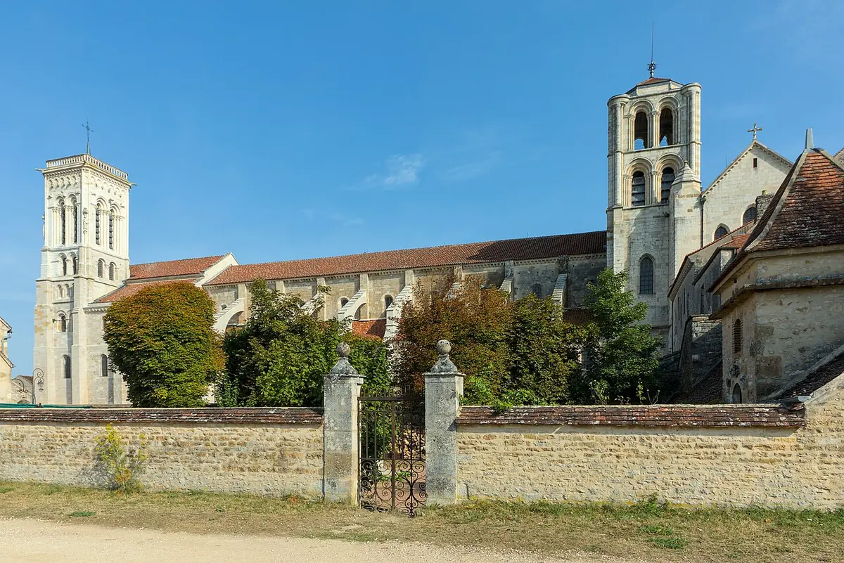 A long Romanesque church with two stone towers on a hilltop, seen across a stone wall and a garden in bright sunshine