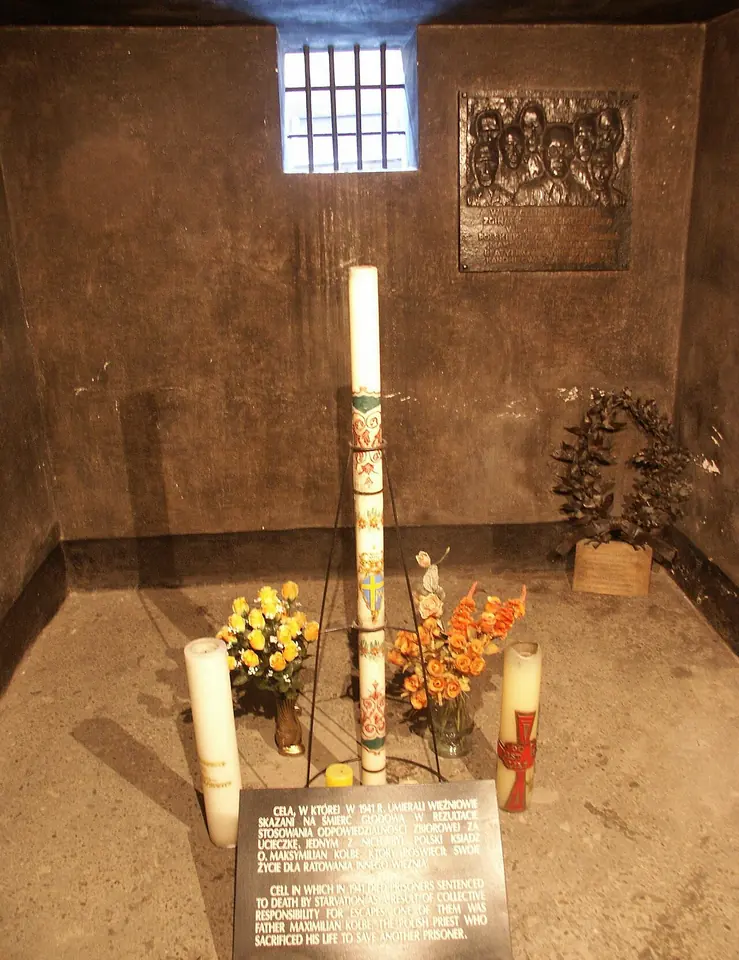 Celda de sótano desnuda con una pequeña ventana enrejada, un cirio pascual alto, flores y una placa conmemorativa en el suelo