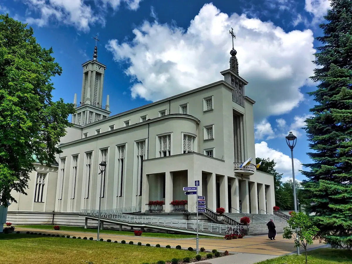 Basílica blanca de estilo moderno con un alto pórtico de columnas y una torre esbelta coronada por una cruz, bajo un cielo azul de verano